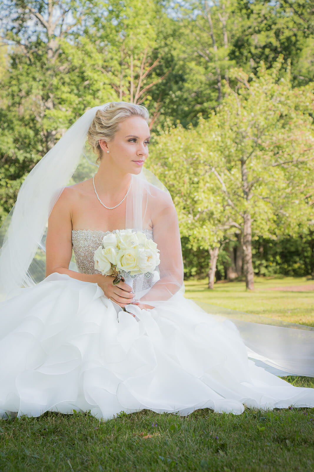 a beautiful bride sitting down near the ground holding her white rose bouquet, looking off into the horizon