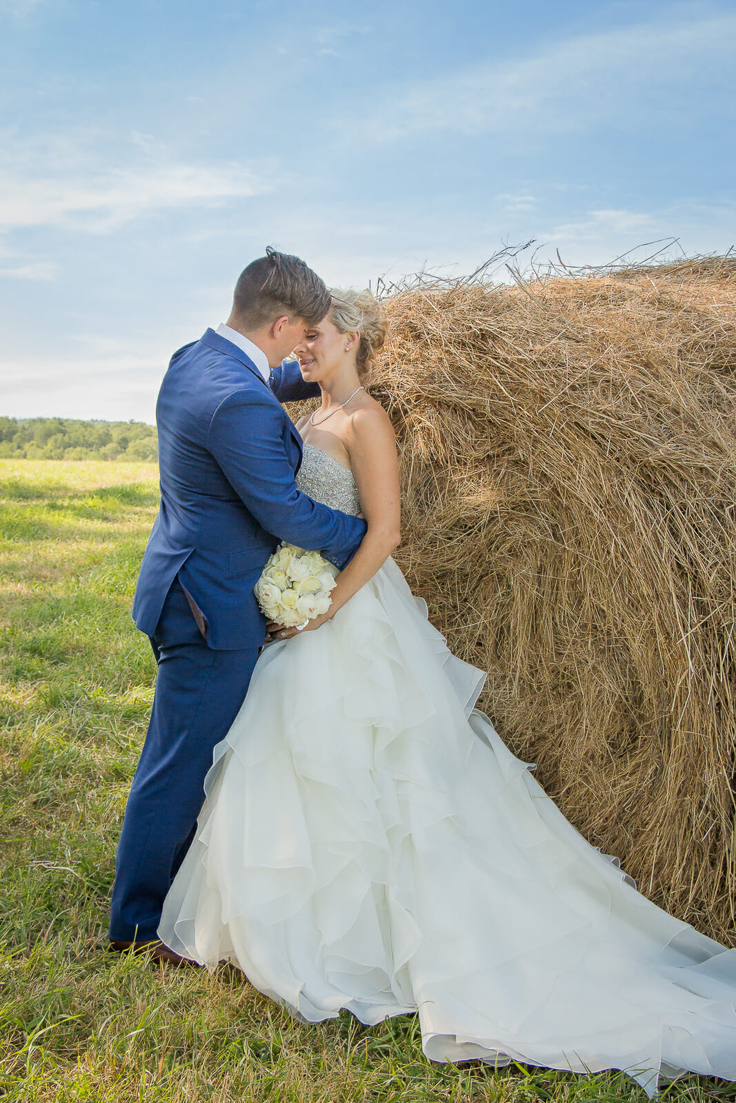 a bride and groom embrace, round bales in the background with a beautiful blue sky.